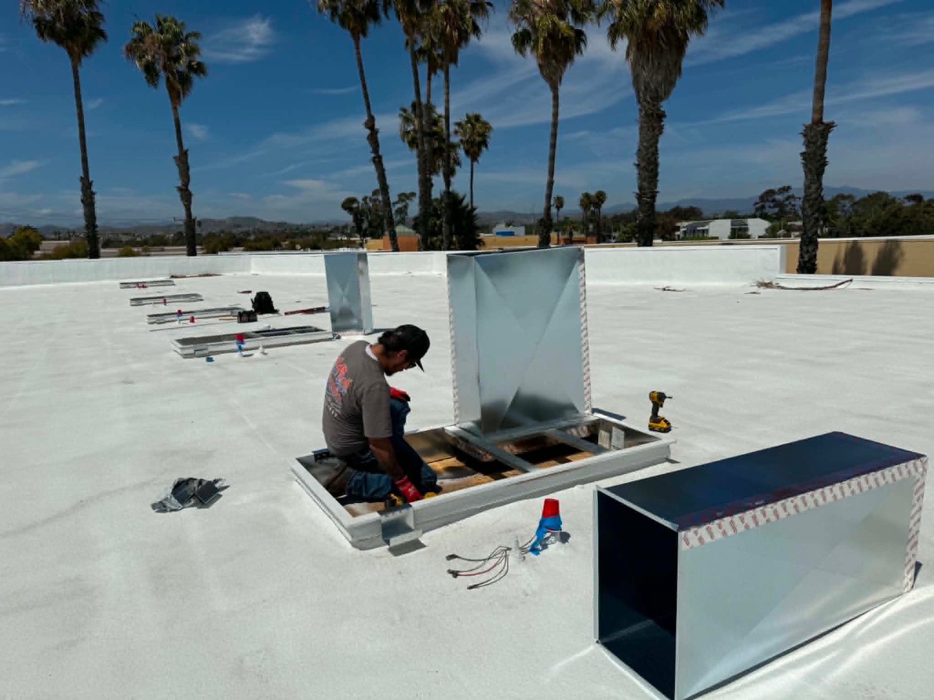 ColdTech technician installing new sheet-metal ductwork on a commercial white flat roof surrounded by palm trees
