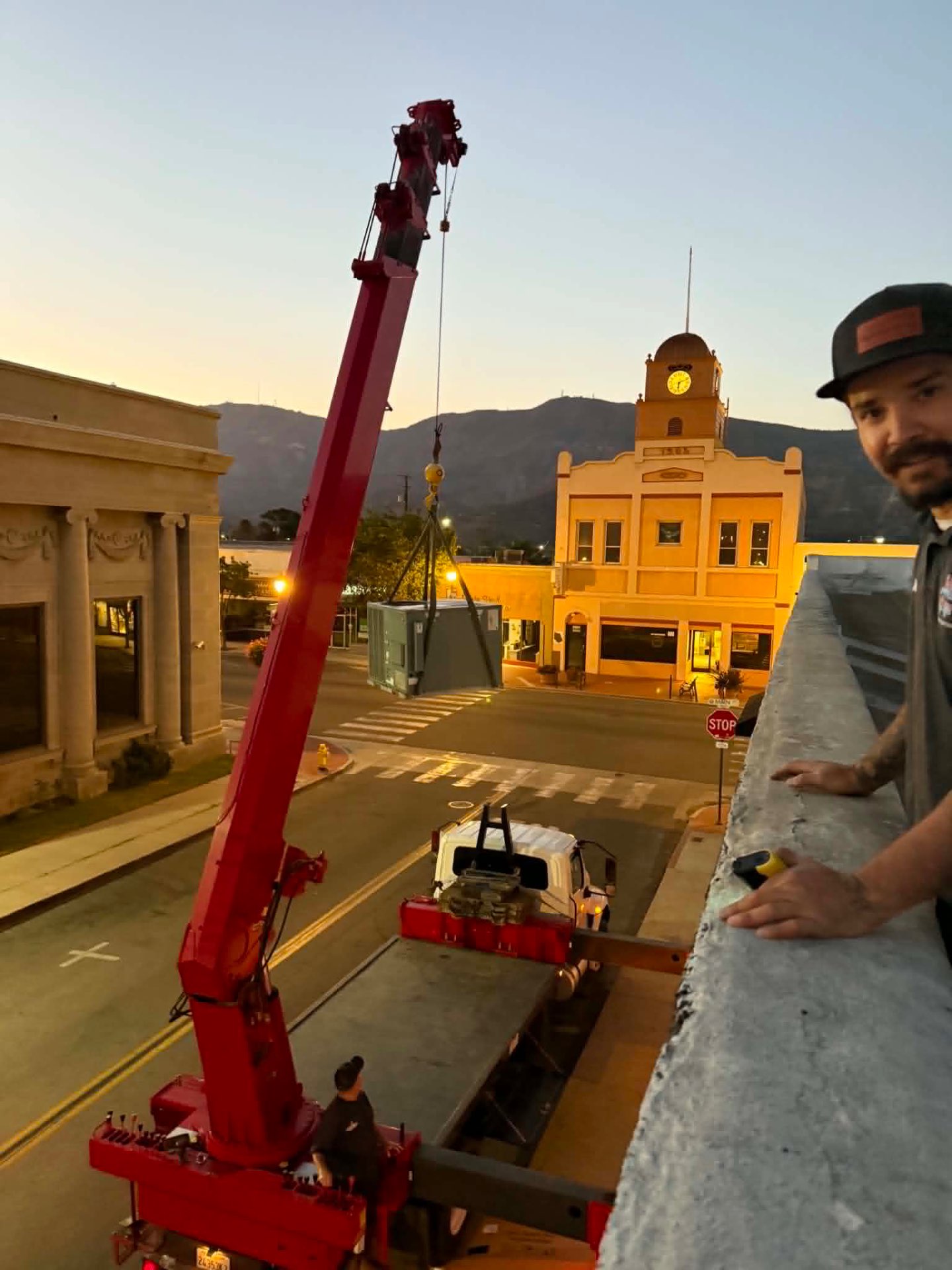 Red boom crane lifting a commercial HVAC unit over a downtown street at sunset with a clock tower in the background